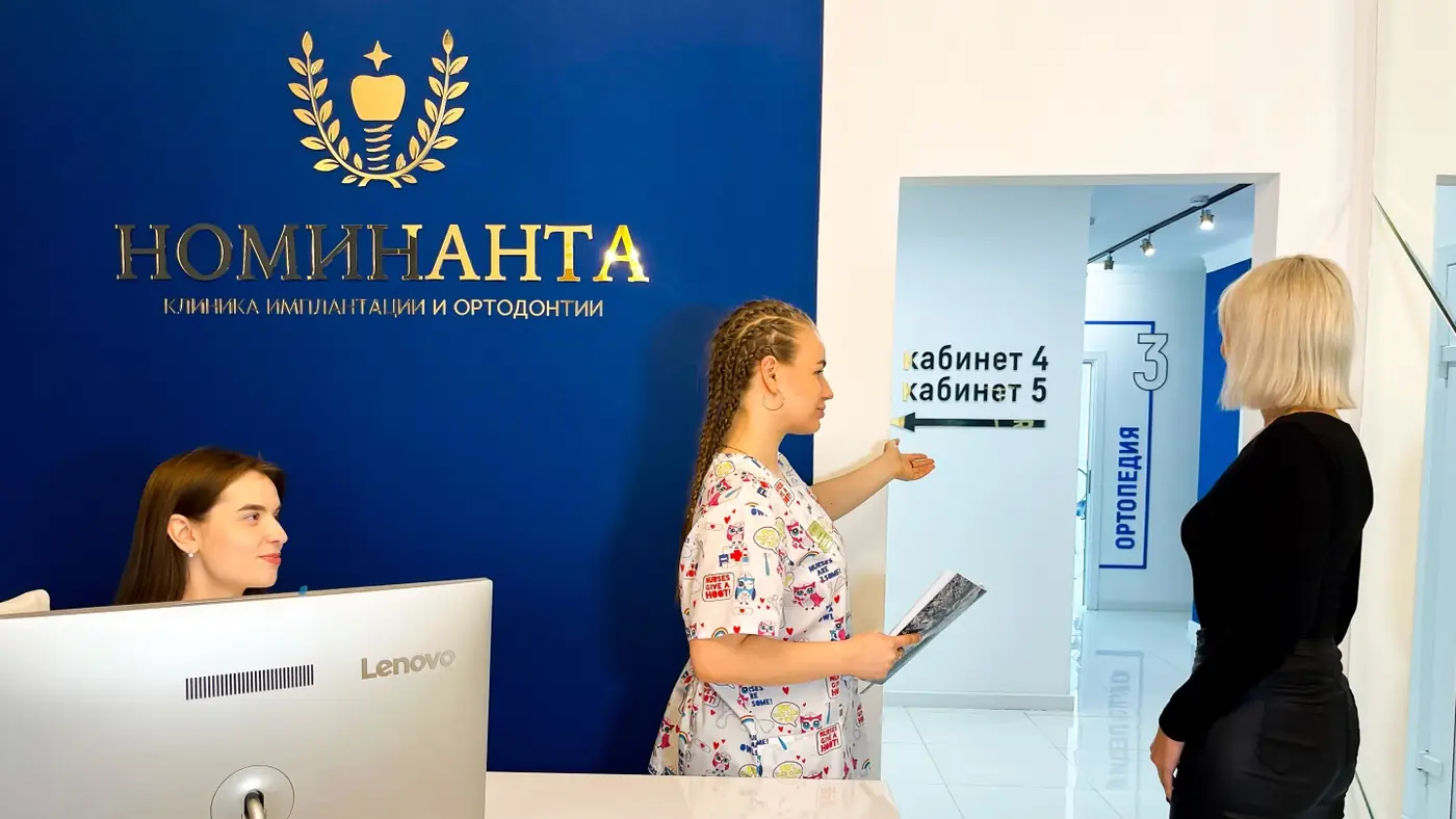Администратор и врач разговаривают с пациенткой на ресепшене клиники «Номинанта»